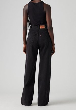 Person with long braided hair wearing a black sleeveless top and loose-fitting high-waisted black jeans, facing away against a gray background.