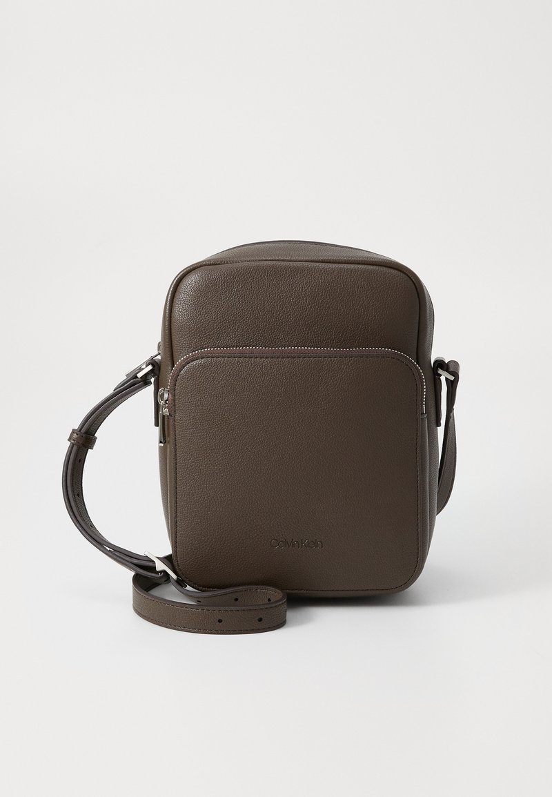 Brun skinn crossbody-veske med teksturert overflate, rektangulær form, frontlomme med glidelås og justerbar stropp. Logo vist på forsiden.