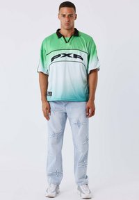 Un uomo sta in piedi di fronte, indossando una polo a maniche corte con sfumature dal verde al bianco e jeans azzurri chiari con motivi a forma di stella cuciti.
