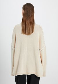 Suéter de punto oversized en color crema con detalle de calados en las mangas y bolsillos. Presenta un corte relajado y una textura suave.
