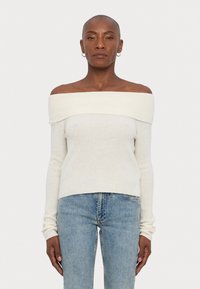 Krema pulover z off-the-shoulder krojem, ribano teksturo, dolgimi rokavi in oprijetim dizajnom, v kombinaciji s svetlo modrimi ravnimi kavbojkami.