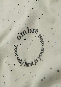 Lichtgrijze stof met een gespikkeld zwart en blauw patroon. Cirkelvormige tekst luidt "jouw enige beperking is je geest" met "ombre" prominent weergegeven.