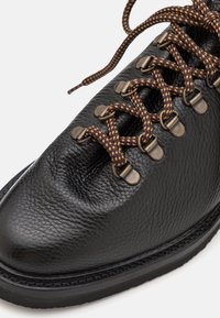 Botte en cuir noir avec une finition texturée, dotée de lacets tressés marron et noir, d'œillets en métal et d'une semelle en caoutchouc noir contrastante.