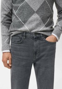 Suéter de punto gris con patrón de diamantes sobre vaqueros de denim gris oscuro. Los vaqueros tienen bolsillos delanteros y un cierre de botón.