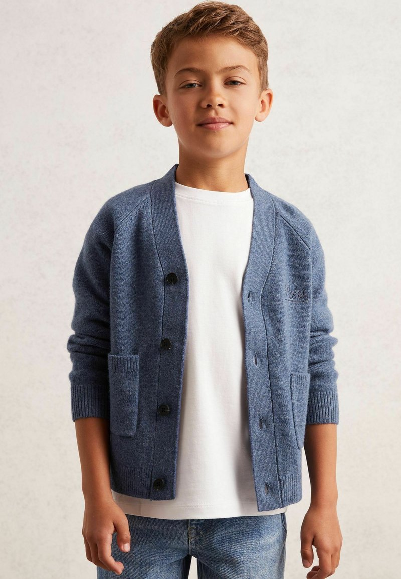 Cardigan blu lavorato a maglia con scollatura a V, chiusura con bottoni, polsini a costine e due tasche frontali, indossato sopra una camicia bianca e jeans blu.