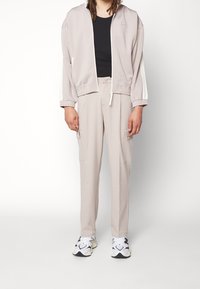 Ljust beige zip-jacket med elastiska manschetter, matchande byxor med cargo-fickor. Svart åtsittande skjorta under; vita sneakers.