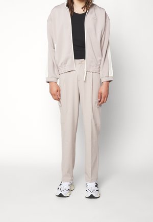 Giacca leggera beige con zip e polsini elastici, abbinata a pantaloni coordinati con tasche cargo. Maglietta nera attillata sotto; sneaker bianche.