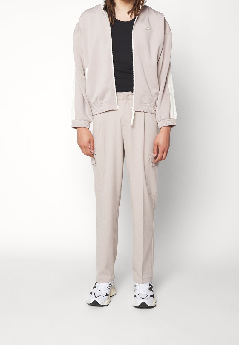 Ljust beige zip-jacket med elastiska manschetter, matchande byxor med cargo-fickor. Svart åtsittande skjorta under; vita sneakers.