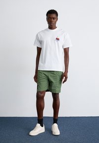 T-shirt bianca in cotone con grafica di ciliegie, abbinata a pantaloni corti verdi in un tessuto leggero, e sneakers bianche con calzini neri.