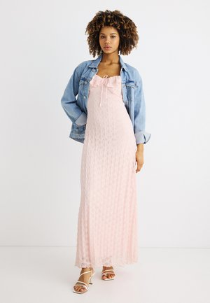 Frau mit lockigem Haar, die ein hellrosa strukturiertes Maxikleid, eine Jeansjacke und weiße Riemchenabsätze trägt, steht vor einem einfarbigen Hintergrund.