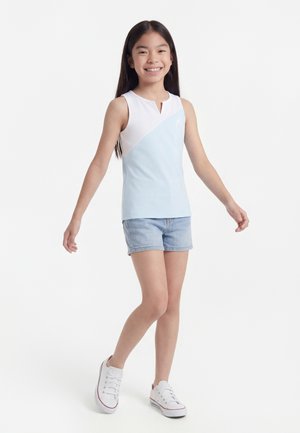 Nasmejana deklica z dolgimi temnimi lasmi, oblečena v brezrokavnik bele in svetlo modre barve, jeans kratke hlače in bele superge, stoji z rahlo razprtimi rokami.