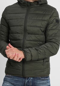 Doudoune vert olive à coupe ajustée, motif matelassé horizontal, fermeture éclair avant et capuche ajustable. Tissu texturé avec finition mate.