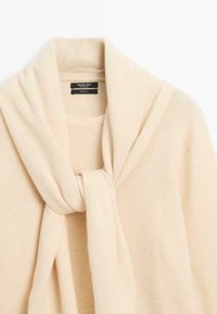 Beige uldsweater med et omslagssnørredesign ved kraven. Har en blød tekstur og en klassisk rund halsudskæring.