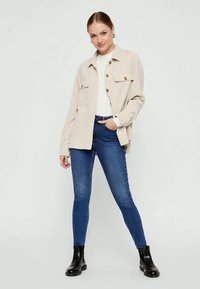 Beige button-up jacket with two chest pockets, worn over a white top and blue skinny jeans, paired with black ankle boots.