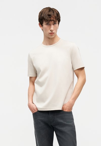 Νέος άνδρας με καστανά μαλλιά, φορώντας μια απλή μπεζ κοντομάνικη μπλούζα και σκοτεινό τζιν, στέκεται με τα χέρια στις τσέπες μπροστά σε λευκό φόντο.