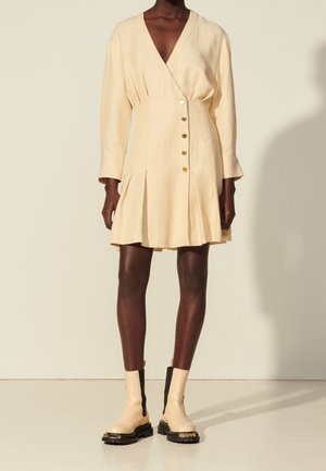 Donna che indossa un vestito crema con bottoni e pieghe, abbinato a stivaletti beige con lati elastici neri e suole spesse.