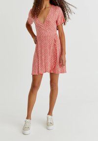 Robe portefeuille rouge à fleurs avec des manches courtes, décolleté en V, détail volanté sur le côté et tissu léger, associée à des baskets beiges.