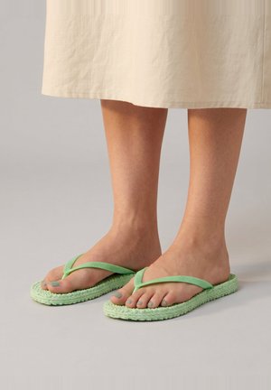 Feet wearing light green braided flip-flops with matching toenail polish, standing on a neutral background beneath a beige skirt.