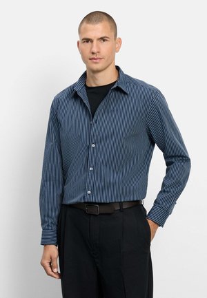 Mann mit kurzen Haaren, der ein blau-weiß gestreiftes Langarmshirt über einem schwarzen Shirt, schwarze Hose und schwarzen Gürtel trägt und mit einer Hand in der Tasche steht.