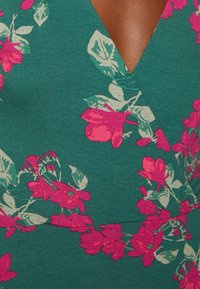 Grön tyg med rosa blommönster, V-ringad design, med stora blommor och blad i ljusare nyanser. Mjuk textur med stretch.