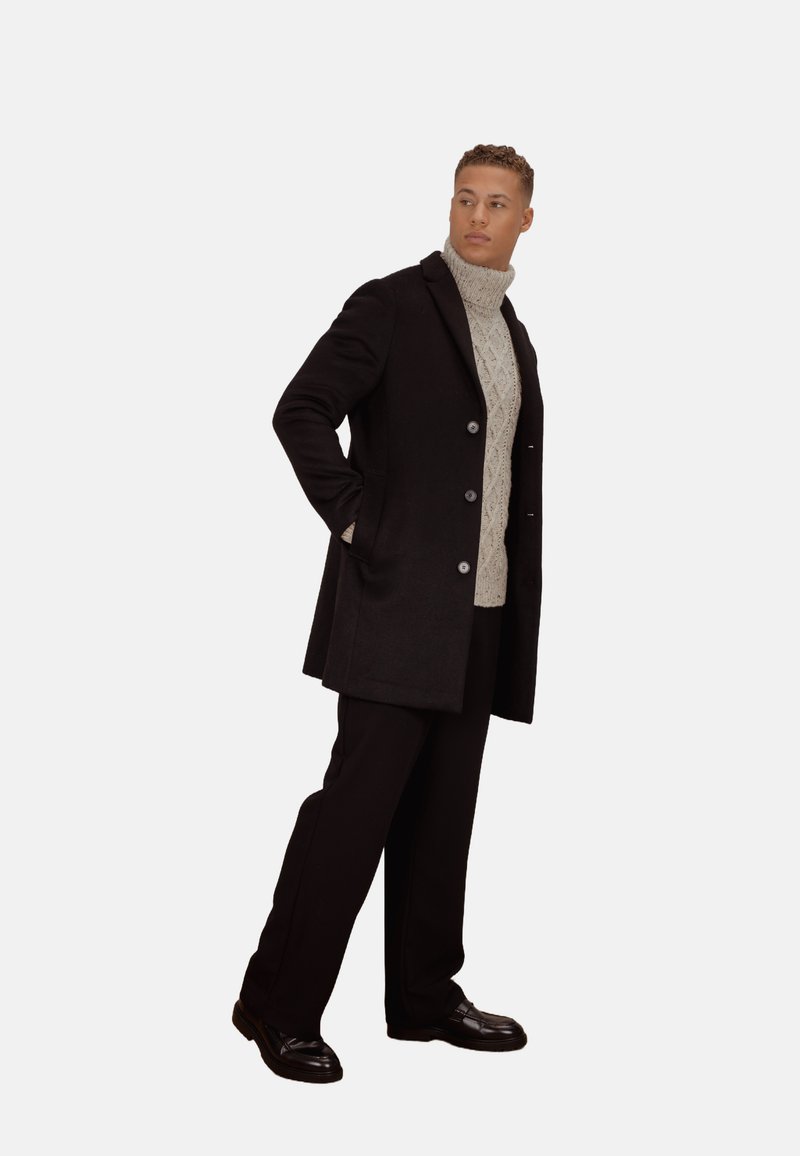 Svart ullfrakk med sølvknapper, kombinert med en kremfarget kabelstrikket høyhalset genser og svarte bukser, båret med skinnende svarte sko.