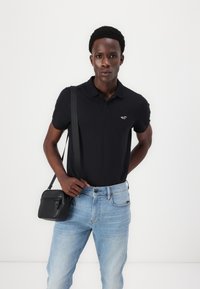 Mlad moški, oblečen v črno polo majico in svetlo modre jeans hlače, nosi majhno črno torbico preko rame.