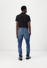 Bărbat în picioare, întors cu spatele, purtând un tricou polo negru, blugi albaștri cu buzunare pe spate și adidași negri cu dungi albe.