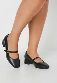 Svarta läder Mary Jane-skor med låg klack, böjd tå och en dekorativ rem med spänne över fotryggen.