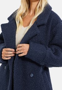 Marineblauer, strukturierter Wollmantel mit einem Reverskragen, übergroßer Passform, zwei Knopfverschlüssen und dezenten Oberflächendetails.