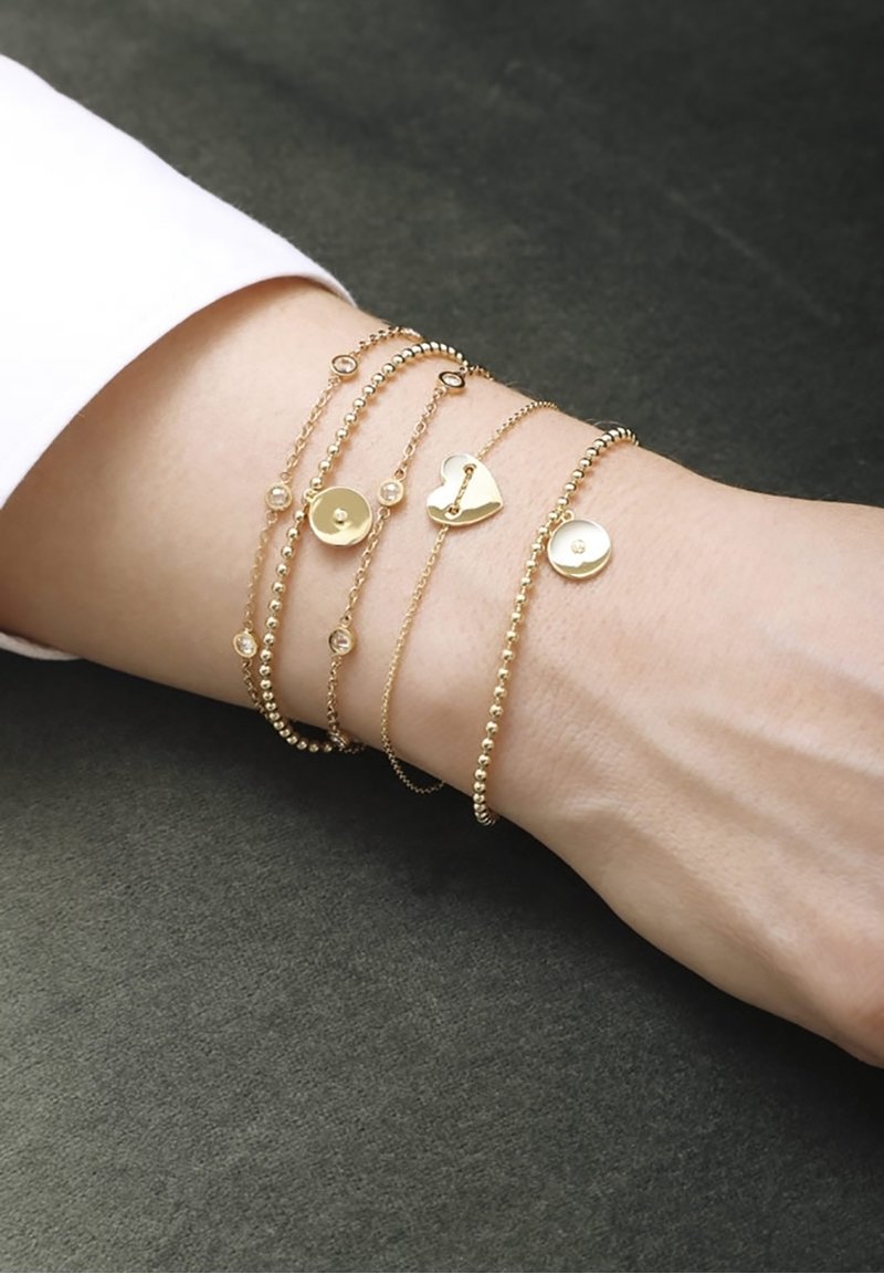 Poignet orné de cinq bracelets délicats en or, présentant de petites perles, des breloques circulaires avec des pierres, et une breloque en forme de cœur avec un motif de serrure.