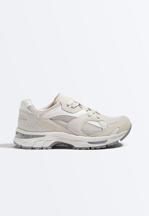 Bézs és fehér sportcipő hálós és velúr betétekkel, fűzős záródással, vastag párnázott talppal és szürke mintázatú talplemezzel.