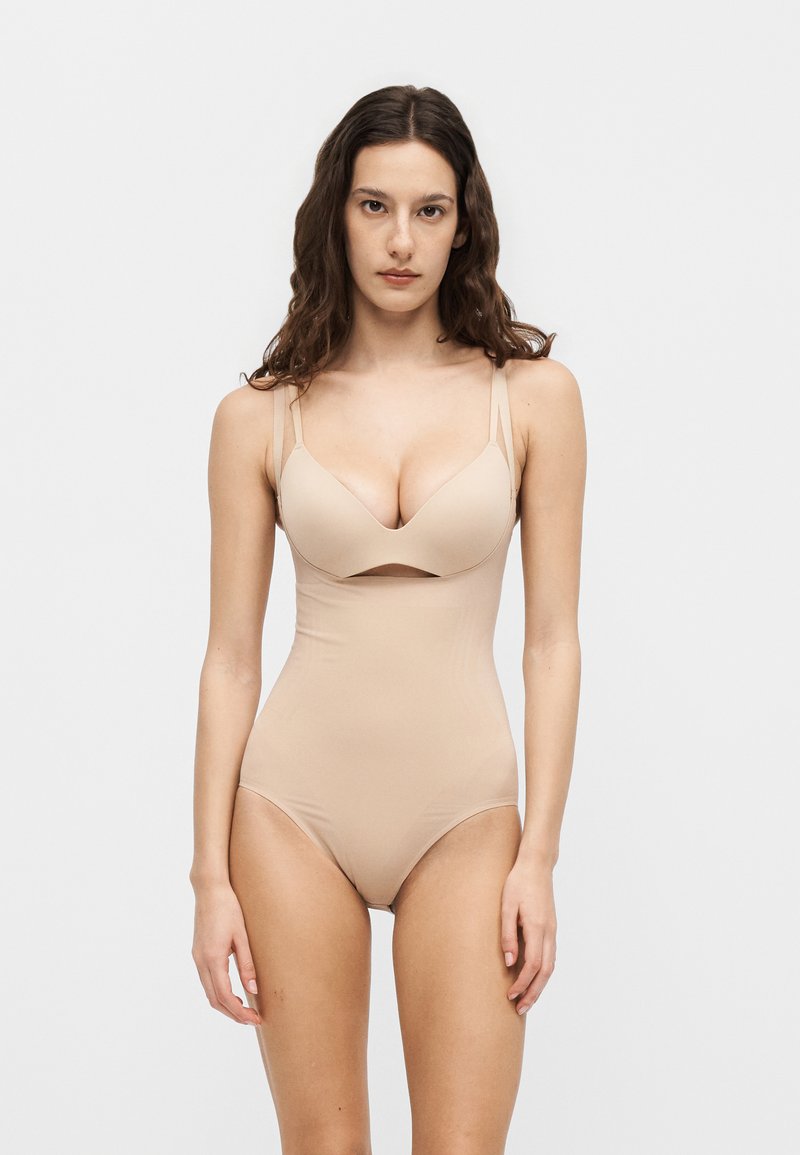 Γυναίκα με μακριά καστανά μαλλιά που φοράει μπεζ αόρατο κορμάκι διαμόρφωσης σώματος, στέκεται μπροστά σε απλό ανοιχτό γκρι φόντο.