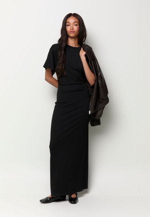 Femme aux longs cheveux foncés portant un haut noir à manches courtes et une jupe longue, tenant une veste en cuir foncé sur une épaule, chaussures plates noires aux pieds.