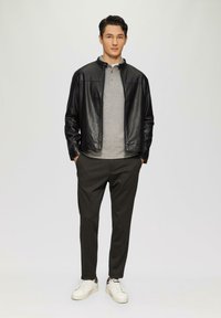 Zwarte leren jack, grijze henley shirt, donkere broek en witte sneakers. Model staat frontaal met handen in de zakken tegen een neutrale achtergrond.
