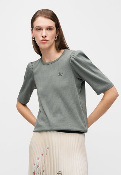 Κοντομάνικο, ριμπ μπλουζάκι σε ελιά χρώμα με στρογγυλή λαιμόκοψη και διακριτικό κεντημένο λογότυπο στο στήθος, συνδυασμένο με πλισέ φούστα.
