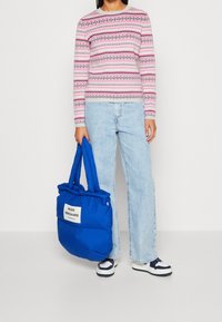 Pull en tricot rose avec des motifs géométriques ; jeans en denim bleu clair ; grand sac doudoune bleu avec étiquette blanche ; baskets marines et blanches.