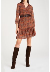 Ruisukuvioinen ruskea mekko, jossa on V-pääntie ja röyhelöhelma, vyötetty ruskealla vyöllä vyötäröltä. Mallilla on polven ylle ulottuvat ruskeat saappaat.