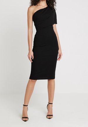 Femme portant une robe midi noire ajustée à une épaule avec un détail à volants et des sandales à talons noires à bride cheville.