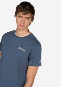 Camiseta de algodón azul con cuello redondo, que presenta la palabra "HARPER" en letras multicolores en el pecho y un logo en la manga.