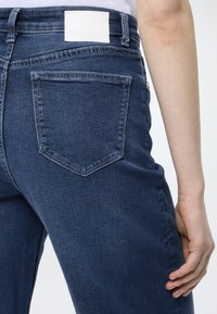 Σκούρο μπλε τζιν παντελόνι με ομαλή υφή, που διαθέτει πίσω τσέπη και λευκή ετικέτα στη μέση. Καθαρός, κλασικός σχεδιασμός.