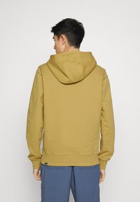 Sweatshirt amarelo mostarda com capuz, de corte relaxado, com punhos e barra canelados. Apresenta uma costura na parte de trás do pescoço e uma pequena etiqueta de logo na parte inferior.