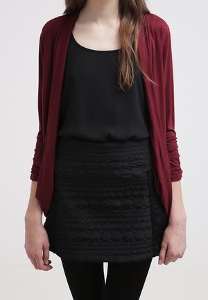 Cardigan drapé bourgogne par-dessus un haut sans manches noir, associé à une jupe noire texturée avec des motifs circulaires en relief, et des collants noirs.