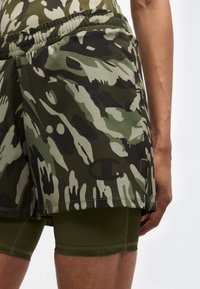 Άτομο που φοράει αθλητικό σορτς με πράσινο καμουφλάζ και μαύρο λογότυπο, με εσωτερικά ελαιώδη σορτς συμπίεσης, στέκεται μπροστά σε λευκό φόντο.