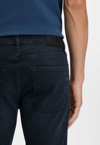 Man med mörkblå jeans med en BOSS-etikett på midjan och en blå skjorta instoppad, arm avslappnat vid sidan.