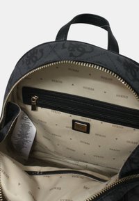 Intérieur du sac à dos à motifs noir et gris avec une poche zippée, présentant un doublage beige clair marqué du mot "GUESS" répétée.
