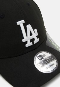 Gorra ajustable negra con el logotipo "LA" bordado en blanco. Presenta una forma redondeada y una tela texturizada. Incluye una etiqueta redonda plateada etiquetada como "9FORTY".