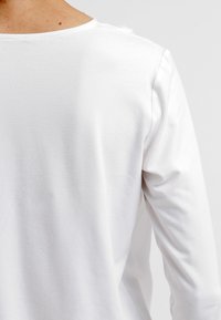 Chemise blanche à manches longues en coton doux, avec une texture lisse et un col rond, dotée de coutures propres au niveau des épaules.