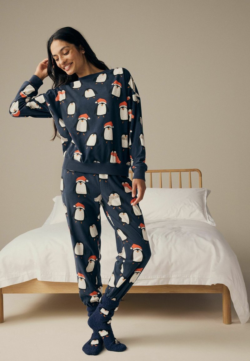 Marineblaues Pyjama-Set mit Pinguinmustern, die rote Weihnachtsmützen tragen. Weicher Stoff, lange Ärmel, elastischer Taillenbund und passende Socken.