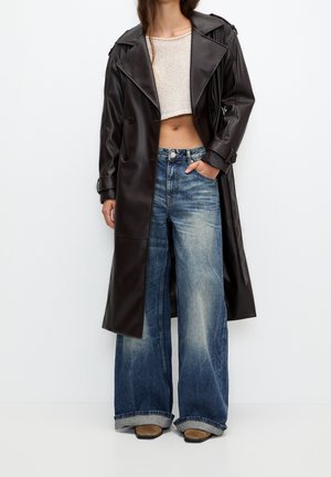 Kvinde iført en lang mørkebrun læderfrakke, kort beige striktop, viddebenede, falmede blå jeans og tanfarvede spidse sko, stående mod en hvid baggrund.