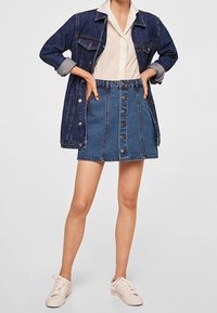Giacca di jeans blu scuro, con bottoni, maniche arrotolate. Camicia bianca con colletto sotto. Gonna di jeans blu chiaro con bottoni frontali. Sneakers bianche.